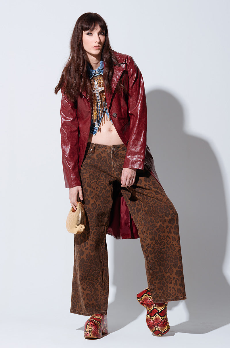 A woman with long brown hair is wearing a red leather coat and the CHEETAH GIRL PANT, which features an oversized wide-leg style in leopard print. Her multicolored top complements the chunky red shoes, while she holds a small gold purse, posing confidently against a plain background.