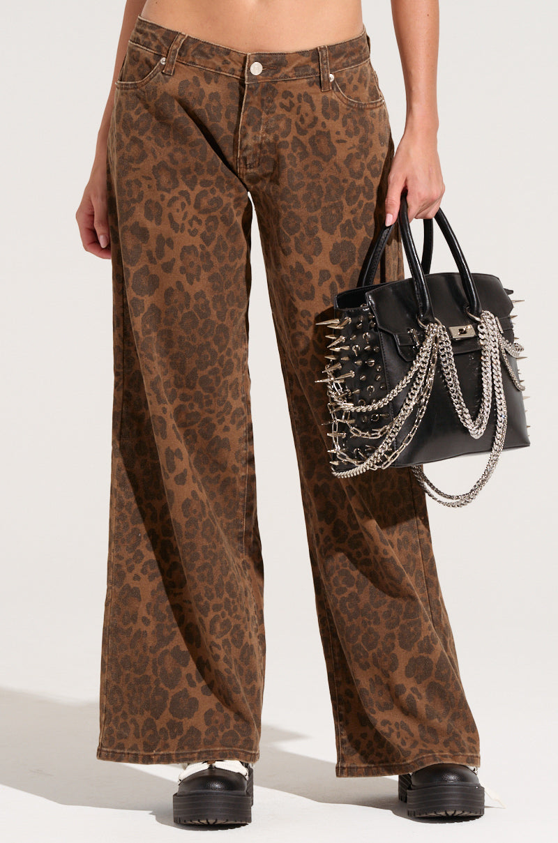 A person is wearing the CHEETAH GIRL PANT featuring a brown, high-rise waist with a wide-leg silhouette and black platform shoes, holding a black handbag embellished with metal spikes and chains. The image highlights the lower half, emphasizing the intricate cheetah pattern and bold accessories.