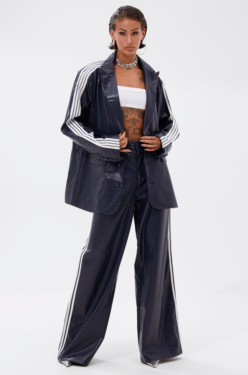A person stands confidently in the DAYTIME LOVIN TROUSER IN NAVY, paired with a matching jacket, white bandeau top, chunky necklace, slicked-back hair, and visible torso tattoos.