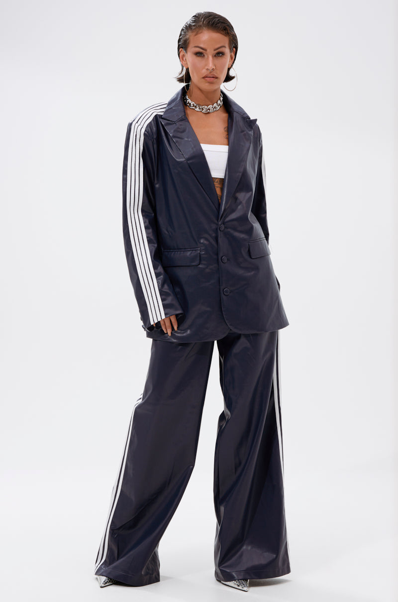 A woman stands against a plain background wearing the DAYTIME LOVIN TROUSER IN NAVY, a high-rise wide-leg pant with athletic stripes, paired with a white bandeau top, silver heels, and a chunky silver necklace.