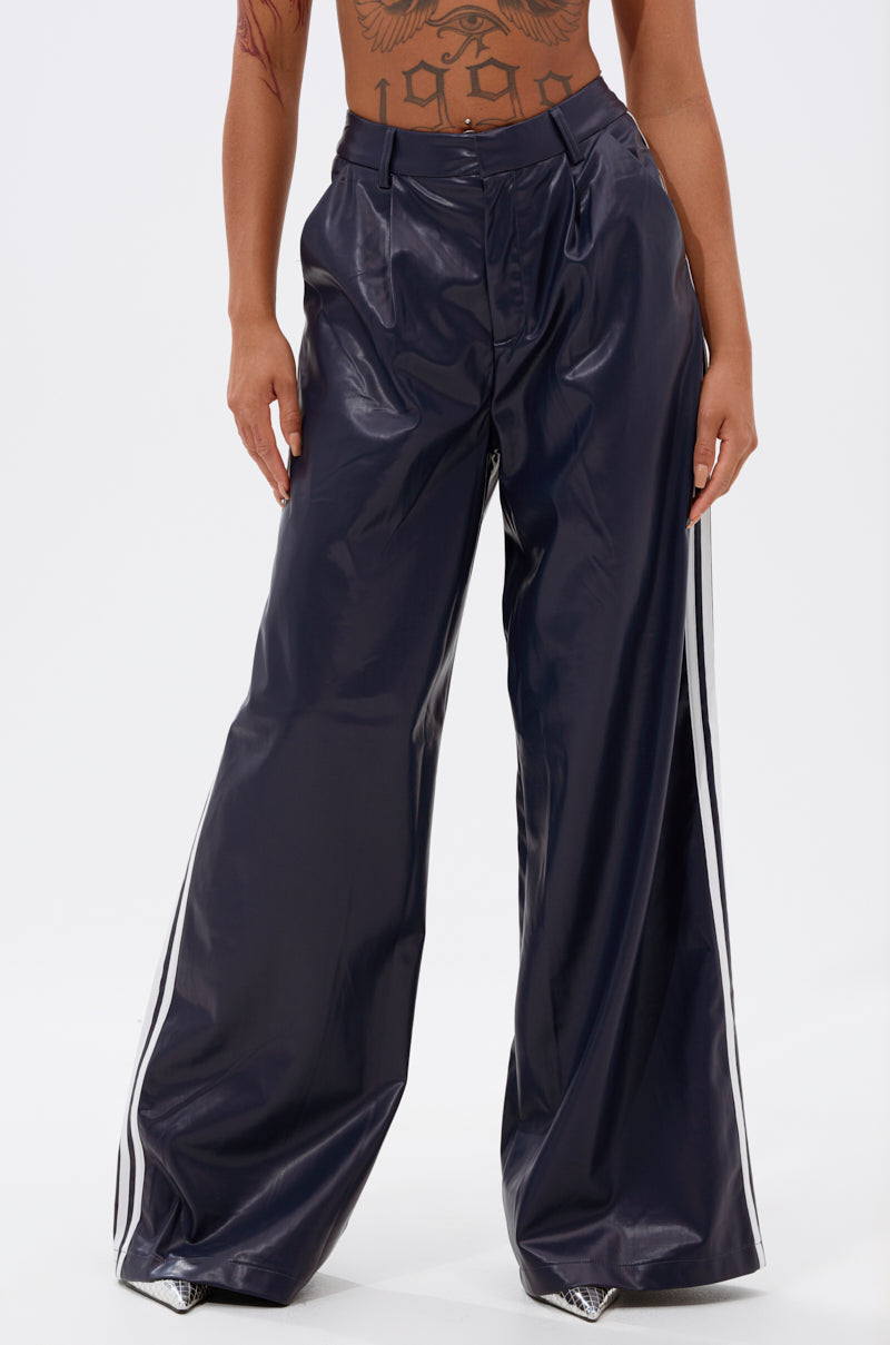 Someone stands against a plain white background wearing the DAYTIME LOVIN TROUSER IN NAVY—shiny, high-rise wide-leg pants with white side stripes, showcasing their midriff and tattooed torso.