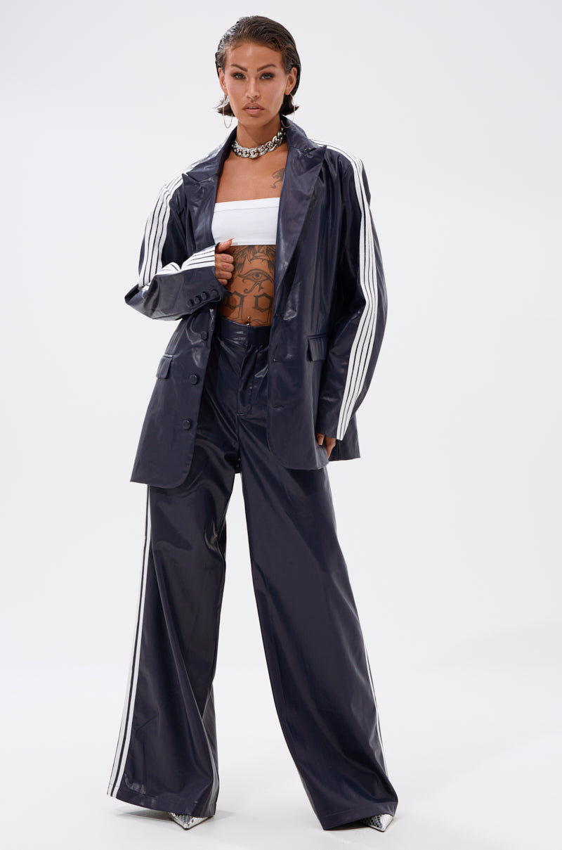 A person stands confidently in the DAYTIME LOVIN TROUSER IN NAVY with white stripes, paired with a matching jacket, white bandeau top, chunky necklace, and visible tattoos, against a plain white background.
