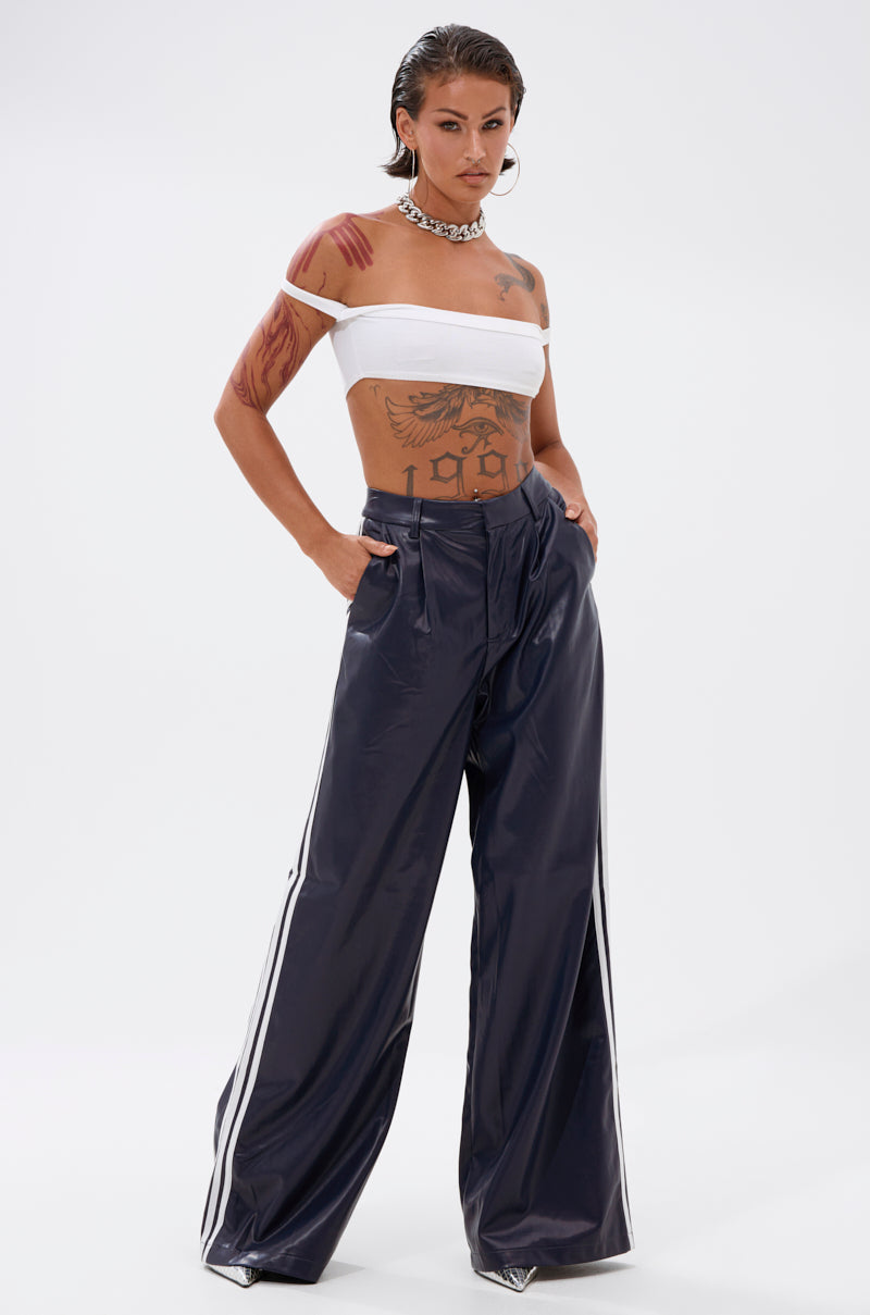 A person with slicked-back hair wears a white off-shoulder crop top, the DAYTIME LOVIN TROUSER IN NAVY featuring white athletic stripes, a chunky chain necklace, and has visible tattoos on their arms and torso.