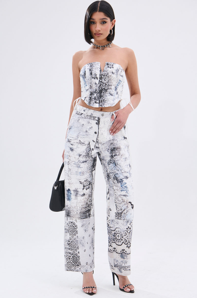 A woman stands against a plain white background wearing the READ ME TROUSER, a high-waisted pant with abstract linework print, paired with a matching strapless AKIRA Label corset top. Her look is styled with black heeled sandals, a black handbag, and a chunky necklace.