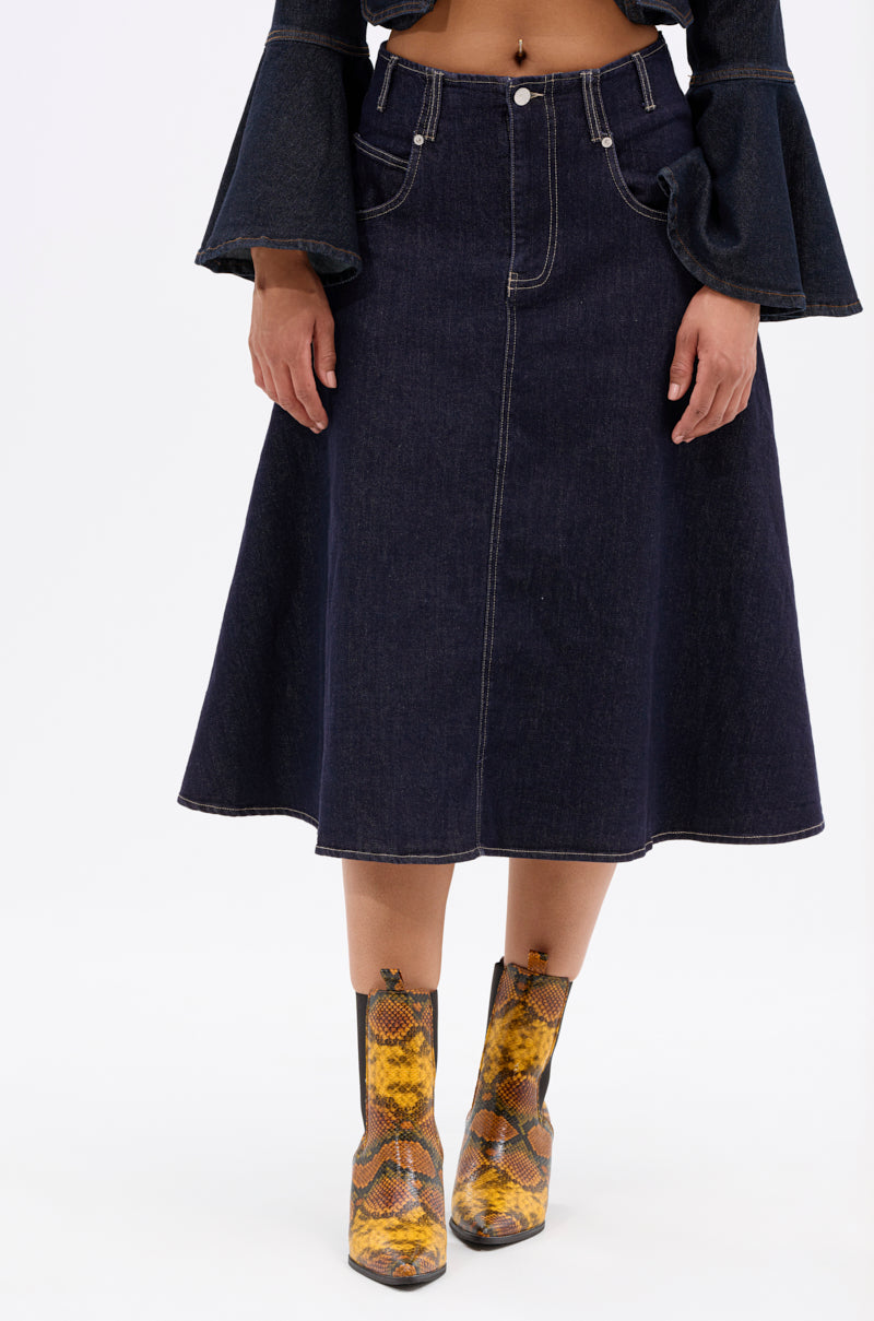 Wearing the COME WITH ME DENIM MIDI SKIRT, a dark long-sleeve top with flared cuffs, and yellow snake-print ankle boots, a person stands against a plain white background.