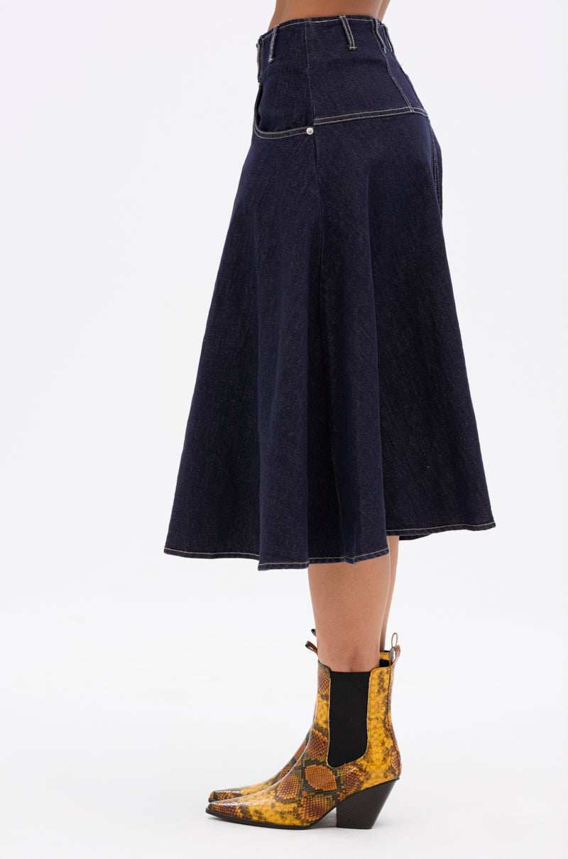 A person wearing the COME WITH ME DENIM MIDI SKIRT, a dark-wash high-rise midi skirt, paired with yellow snakeskin-patterned ankle boots with black elastic side panels, shown in profile against a white background.