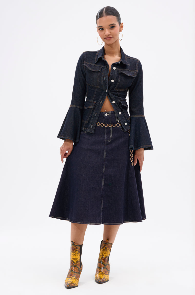 A woman poses in the COME WITH ME DENIM MIDI SKIRT, paired with a matching dark-wash jacket, flared sleeves, chain belt, and yellow patterned ankle boots against a white background.