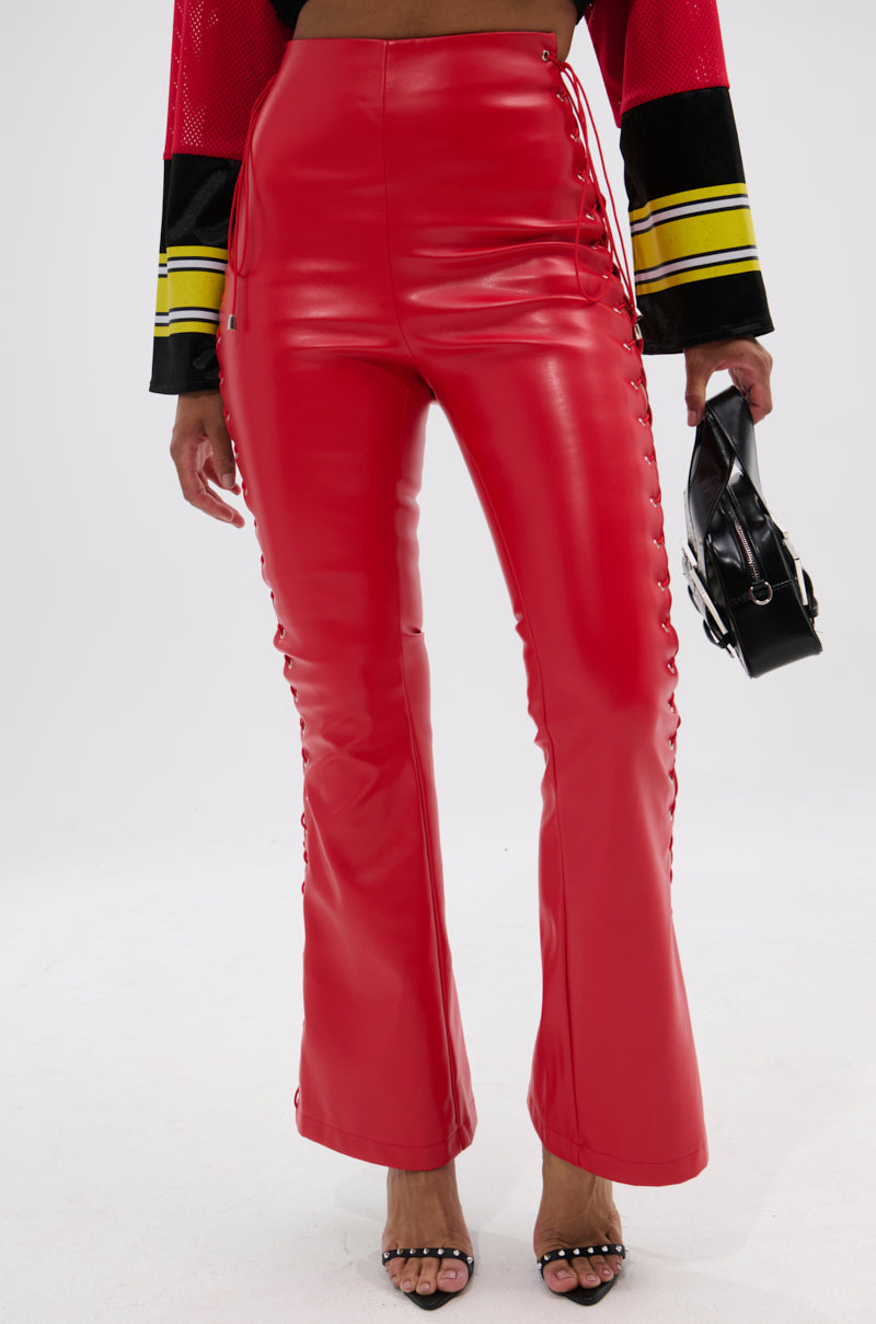 A person in SEXY FAUX LEATHER RED LACE-UP PANTS, a red and black long-sleeve top with yellow stripes, black open-toe heels, and holding a small black handbag stands against a plain white background.