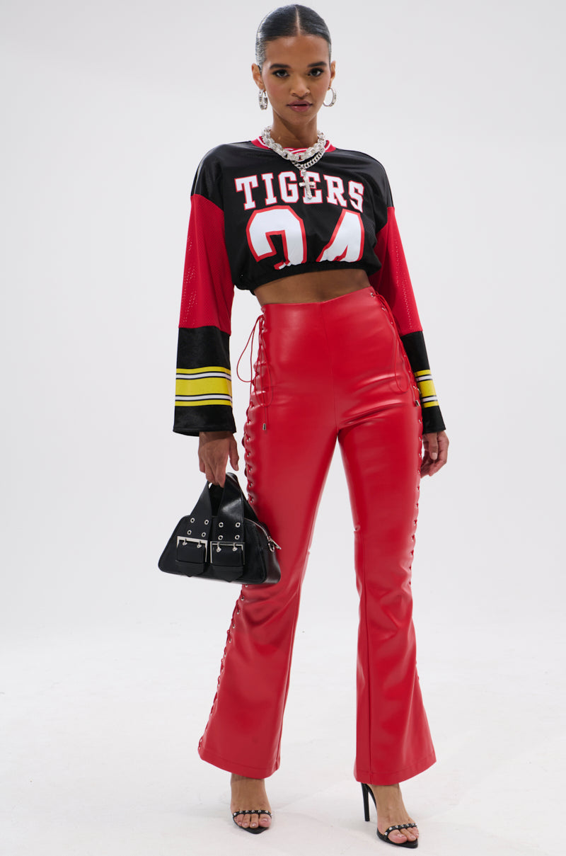 A woman poses in SEXY FAUX LEATHER RED LACE-UP PANTS, a cropped black and red "Tigers 24" jersey, black heeled sandals, and holds a small black handbag against a plain white background.