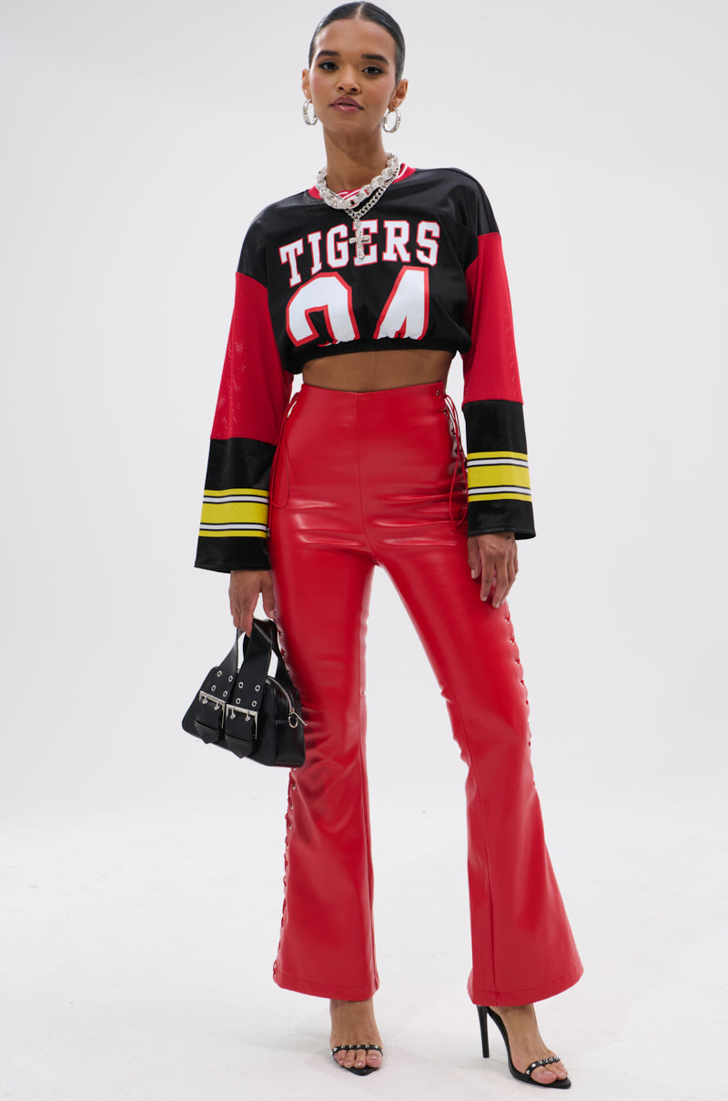 A woman poses in a black and red "TIGERS 24" crop top, SEXY FAUX LEATHER RED LACE-UP PANTS, black strappy heels, and holds a small black handbag against a plain white background.