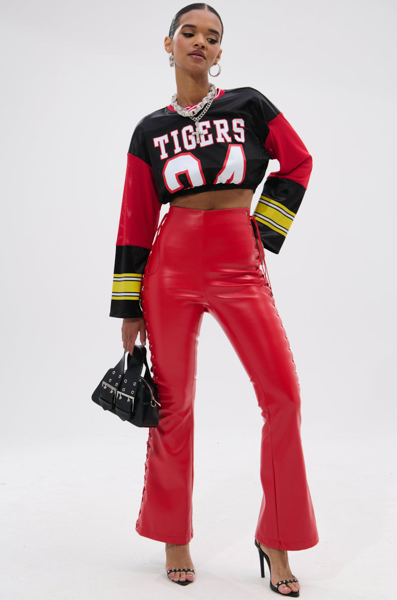 A woman poses confidently in SEXY FAUX LEATHER RED LACE-UP PANTS, paired with a black cropped "Tigers 24" jersey, hoop earrings, necklace, black high-heeled sandals, and a small black handbag.