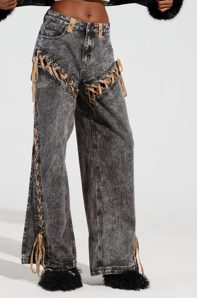 A person is wearing the LACED WITH STYLE LACE UP DENIM JEANS, featuring a high-rise wide-leg cut and acid wash design, with prominent brown lace-up details along the sides and front pockets. These jeans are paired with black furry shoes against a plain white background.