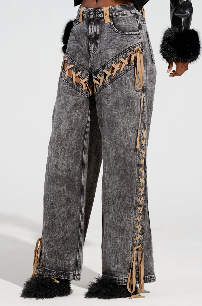 A model showcases the LACED WITH STYLE LACE UP DENIM JEANS in a high-rise, wide-leg gray design featuring tan lace-up detailing on the front and sides, complete with a distressed look. The outfit is paired with a black top featuring fluffy cuffs.
