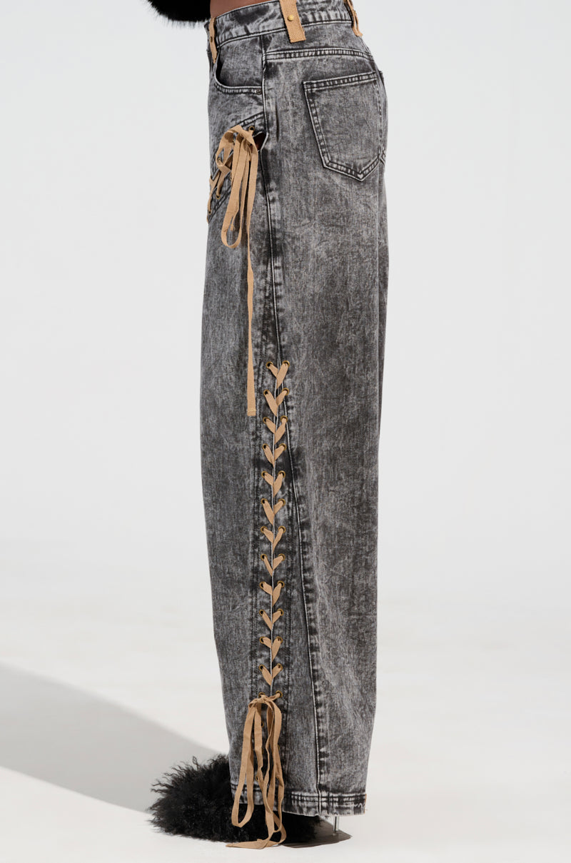 Side view of a person wearing the LACED WITH STYLE LACE UP DENIM JEANS, showcasing high-rise wide-leg gray jeans with intricate lace-up details along the sides. These acid wash jeans feature a washed-out texture and back pockets as the person stands against a plain background.