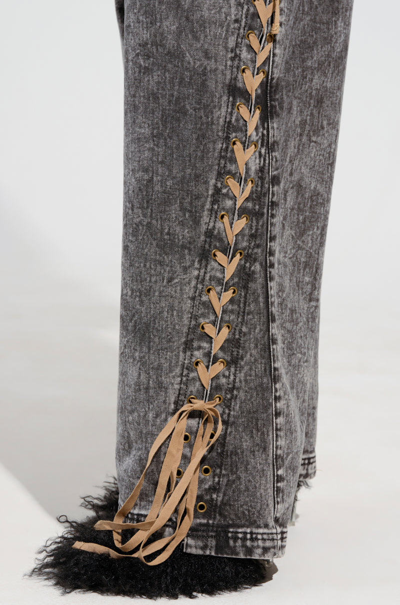 Introducing the LACED WITH STYLE LACE UP DENIM JEANS: a close-up of these faded black, high-rise wide-leg denim pants showcases a distinct tan lace-up design along the side seam. The black fur trim embellishing the hems adds a unique and textured look.