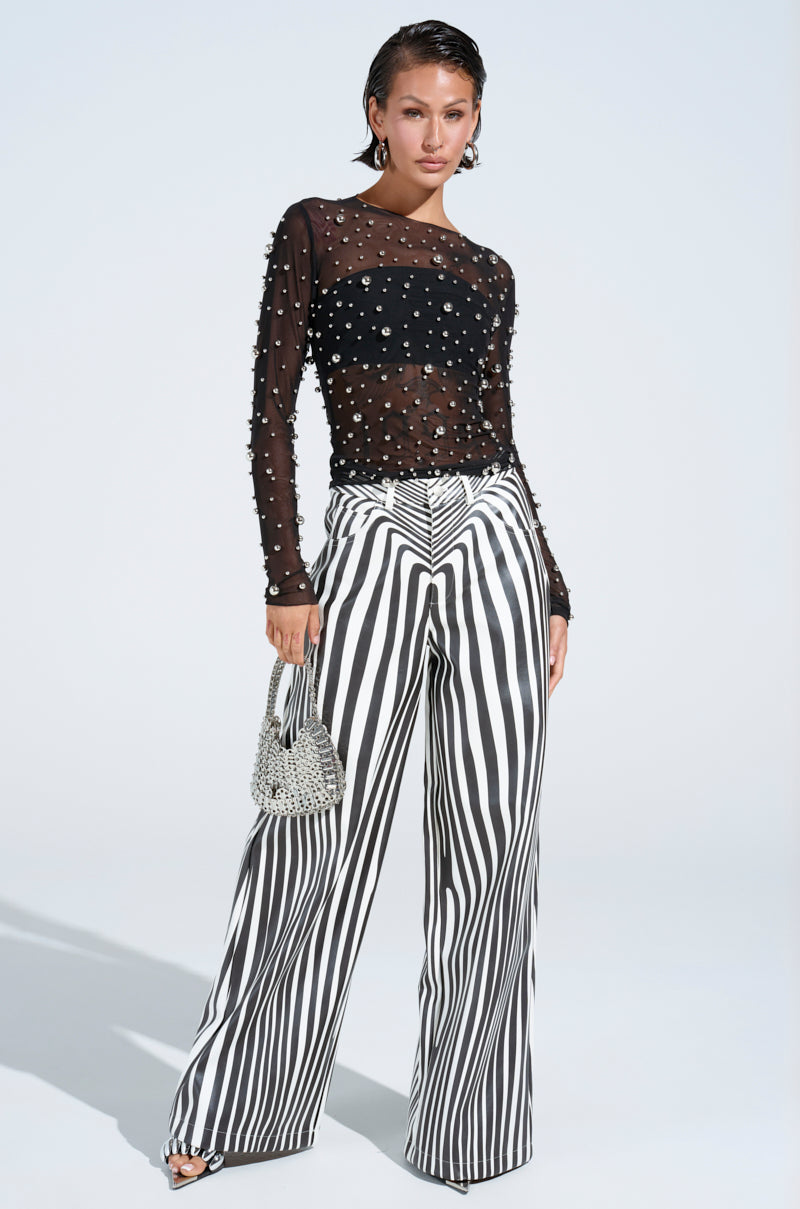 Confidently posing in PARADOX STRIPE PANT, they pair the high-rise fit with a pearl-embellished sheer black top, holding a small beaded handbag and wearing open-toed shoes against a simple light backdrop.