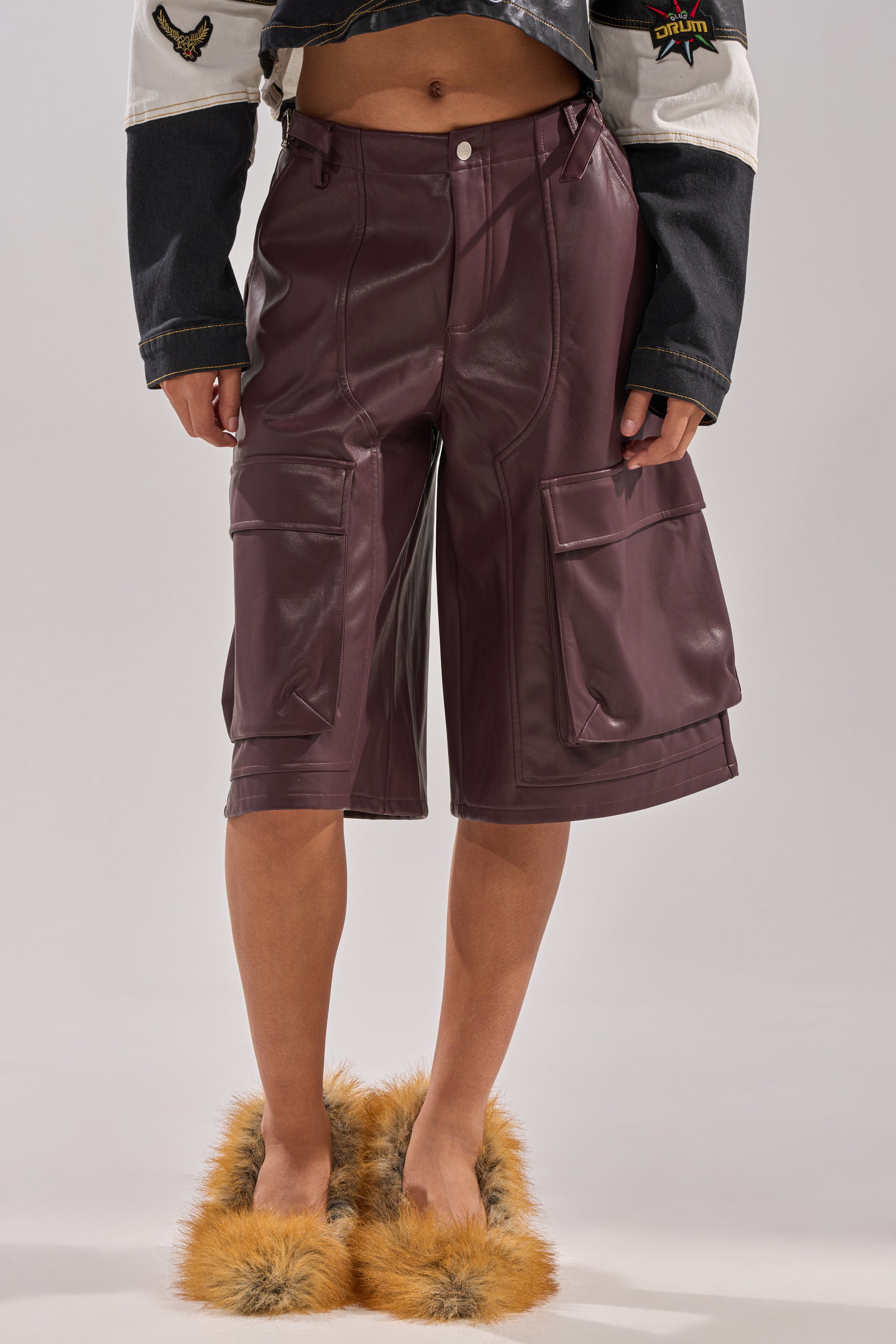 Wearing the OVER DONE FAUX LEATHER BERMUDA SHORT in burgundy with cargo pockets and brown fuzzy slippers, paired with a cropped jacket featuring black sleeves and visible patches. Image is cropped from the chest down.