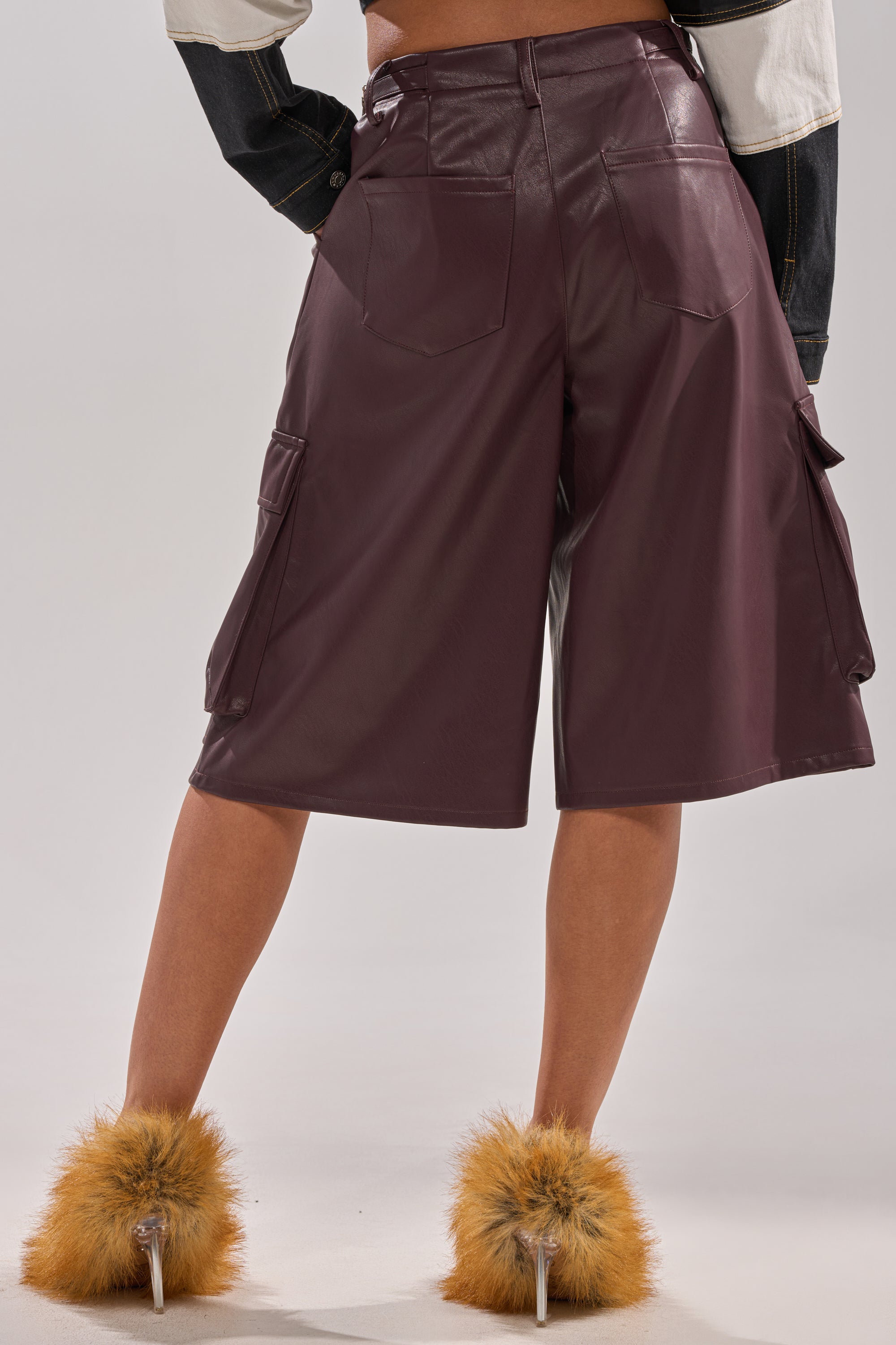 A person stands facing away, wearing the OVER DONE FAUX LEATHER BERMUDA SHORT with large back and side cargo pockets, paired with a cropped jacket and fluffy orange heeled slippers.