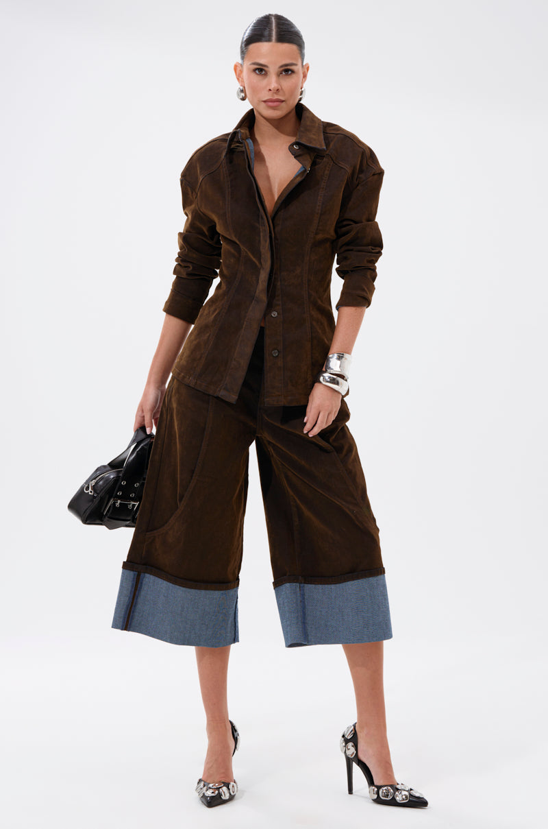 A woman embraces fall fashion in the FALL VIBES VELVET BERMUDA SHORT, styled with a matching brown button-up shirt, denim-cuffed knee-length shorts, a black handbag, pointed heels, silver bracelets, and slicked-back hair.