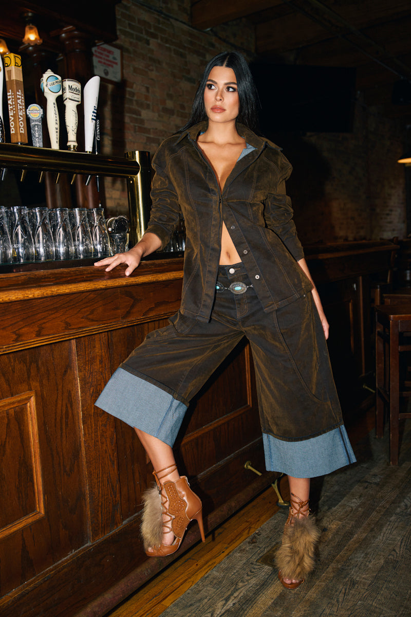 A woman confidently poses by a bar in FALL VIBES VELVET BERMUDA SHORTS, styled with a brown jacket and tan fur-accented heels, embodying chic fall fashion in a dimly lit wooden interior.
