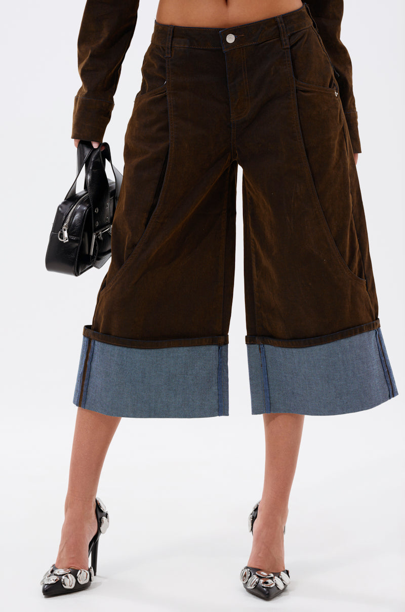 Channeling fall fashion, a person wears the FALL VIBES VELVET BERMUDA SHORT—wide-legged brown culottes with blue denim cuffs—paired with pointed metallic-detailed heels and a small black handbag. Upper body mostly out of frame.