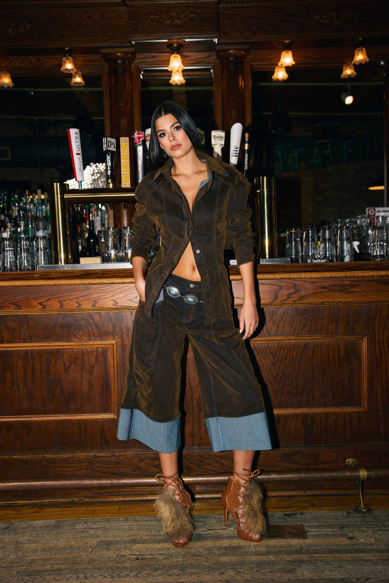 A woman stands by a wooden bar in fall fashion, featuring the FALL VIBES VELVET BERMUDA SHORT, paired with a brown jacket, black top, denim-cuffed pants, belt, and furry brown heeled boots. Glassware and beer taps are behind her.