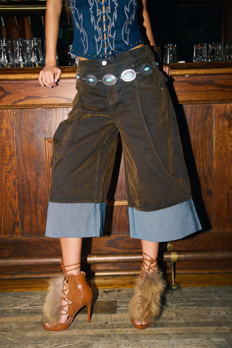 A person in FALL VIBES VELVET BERMUDA SHORTS—brown, wide-leg style with turquoise belt details—paired with a navy lace-up top and tan fur-accented heels, poses before a wooden bar for an effortless fall look.