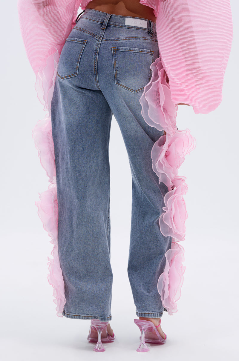 A person wearing FLOWER GIRLIE APPLIQUE DENIM JEANS with pink ruffled sheer side details, a pink textured top, and transparent pink high heels; the photo is taken from behind.
