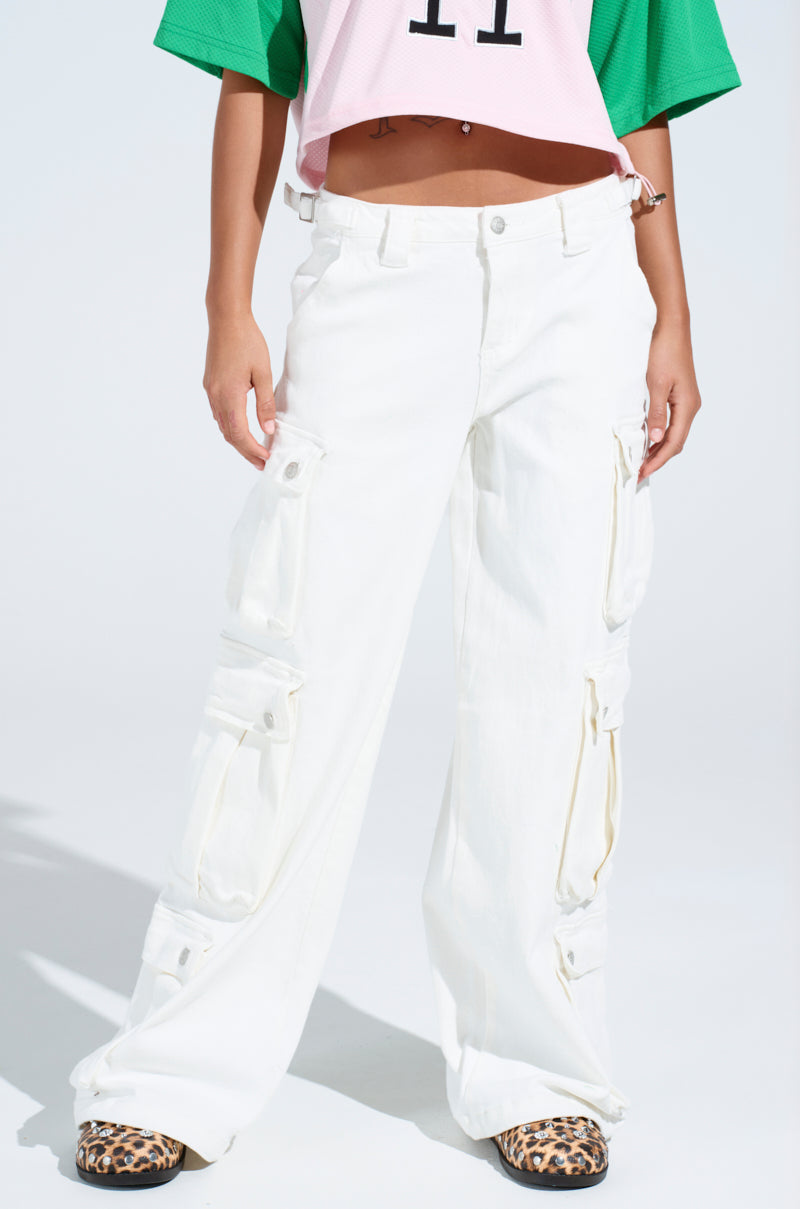 A person wearing a green and pink cropped top with GET RIGHT TO IT CARGO DENIM PANT in a white wash featuring multiple pockets and adjustable buckled straps, paired with leopard print shoes stands against a plain, light-colored background.