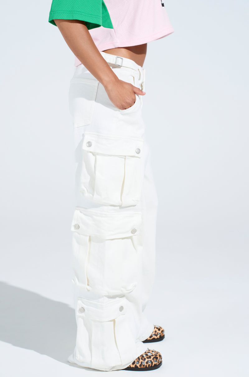 A person dons a green and pink short-sleeved top paired with loose-fitting GET RIGHT TO IT CARGO DENIM PANTS, featuring multiple pockets in a white-wash denim style. The ensemble is finished off with leopard-print shoes, set against a plain white background.