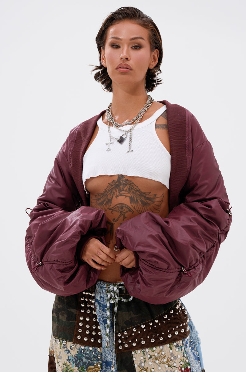 A person with short dark hair wears layered necklaces, a white crop top, patchwork studded pants, and the NO TIME FOR GAMES RUCHED BOLERO maroon jacket. Holding the jacket open, they reveal chest and stomach tattoos.