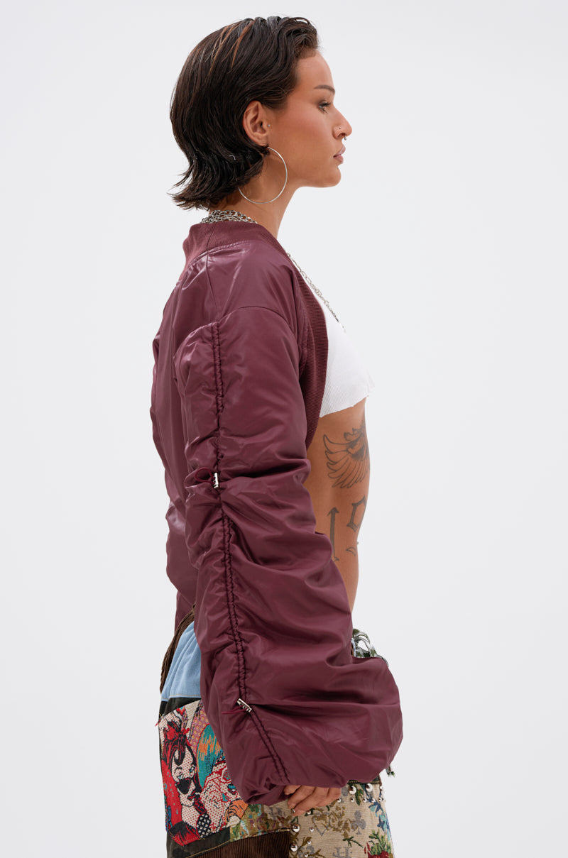 A person with short dark hair and hoop earrings is shown in profile wearing the NO TIME FOR GAMES RUCHED BOLERO in maroon, a white crop top, and patterned pants. Tattoos are visible on their side and arm against a plain light background.