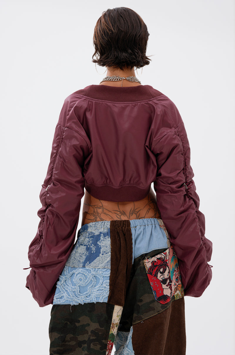 A person with short dark hair, wearing the NO TIME FOR GAMES RUCHED BOLERO in maroon and patchwork pants, stands facing away from the camera against a plain white background.