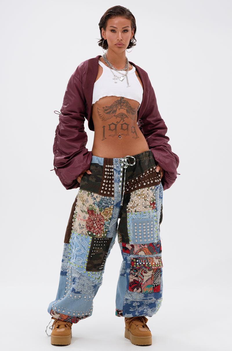 A person with short dark hair wears the NO TIME FOR GAMES RUCHED BOLERO, a white crop top, patchwork baggy jeans with studs, tan boots, and layered necklaces, standing confidently with hands in pockets against a plain white background.