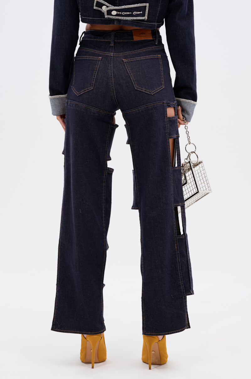 A person stands facing away wearing the LOCKED UP FOR GOOD DENIM PANT, high-rise dark blue jeans with straight legs and large side cut-outs, paired with a matching jacket, tan heels, and holding a boxy handbag.