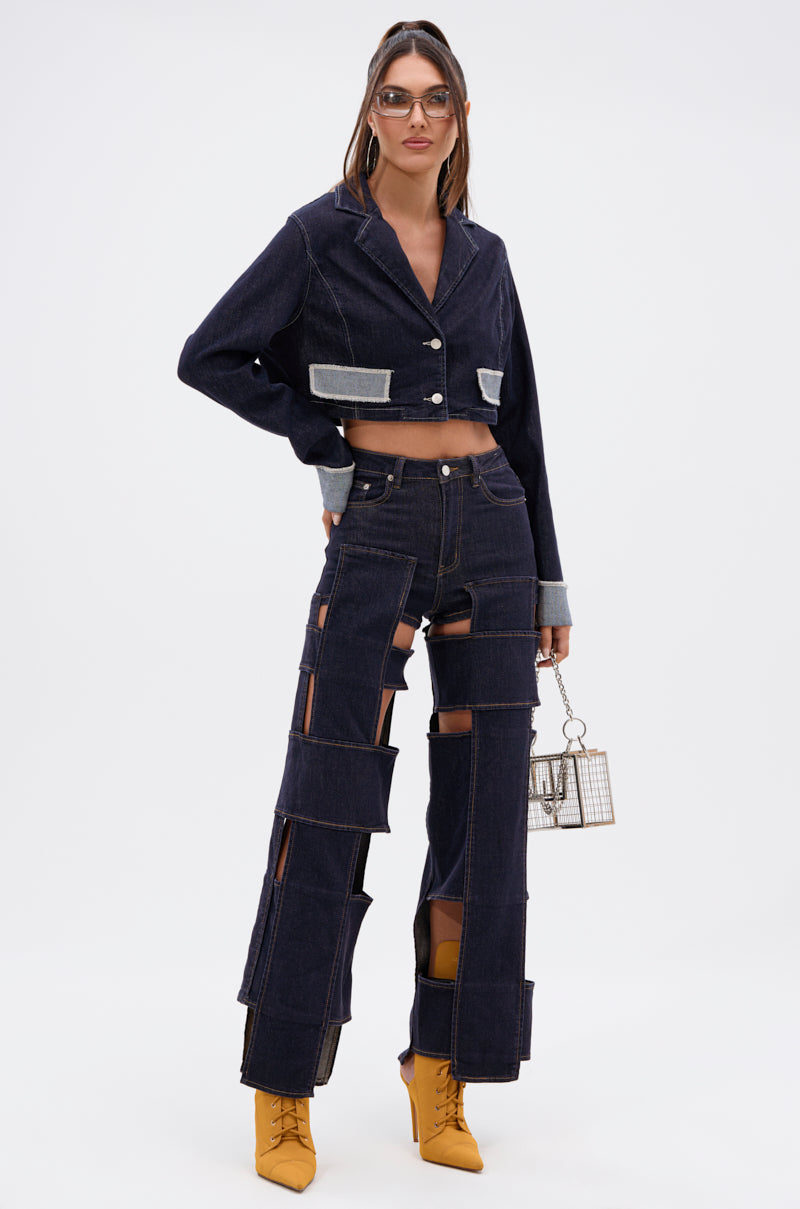A woman wears the LOCKED UP FOR GOOD DENIM PANT, featuring bold cutouts and panels, paired with a cropped denim jacket, yellow heeled boots, sunglasses, hoop earrings, and carries a small cage-style handbag.