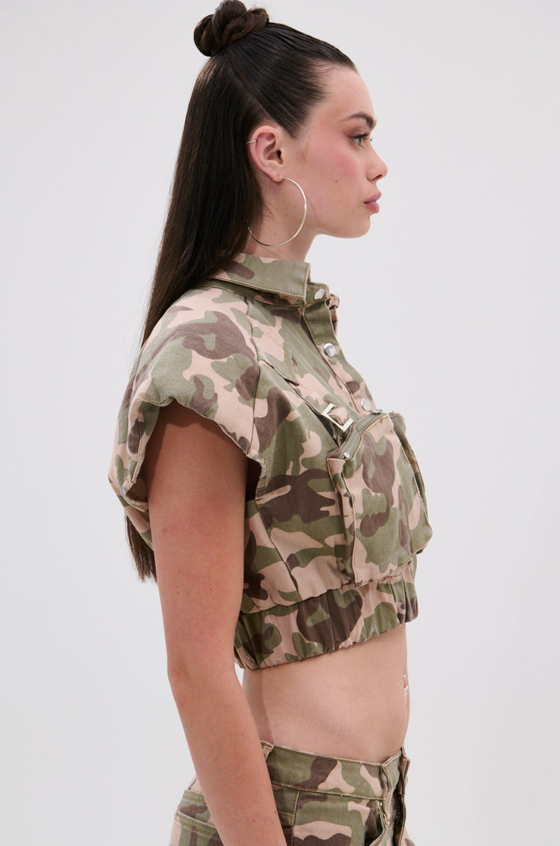 A woman with long dark hair in a half-up top knot models the TAKE ME DOWN CAMO BUTTON DOWN, cropped and paired with matching high-waisted cargo pants, shown in profile against a plain light background.