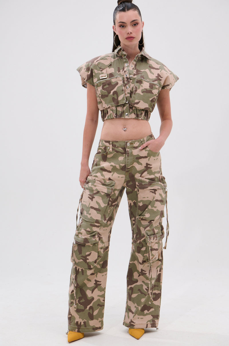 A woman with straight dark hair pulled back stands against a white background wearing the TAKE ME DOWN CAMO BUTTON DOWN, styled as a cropped sleeveless shirt with matching cargo pants and yellow pointed-toe heels.