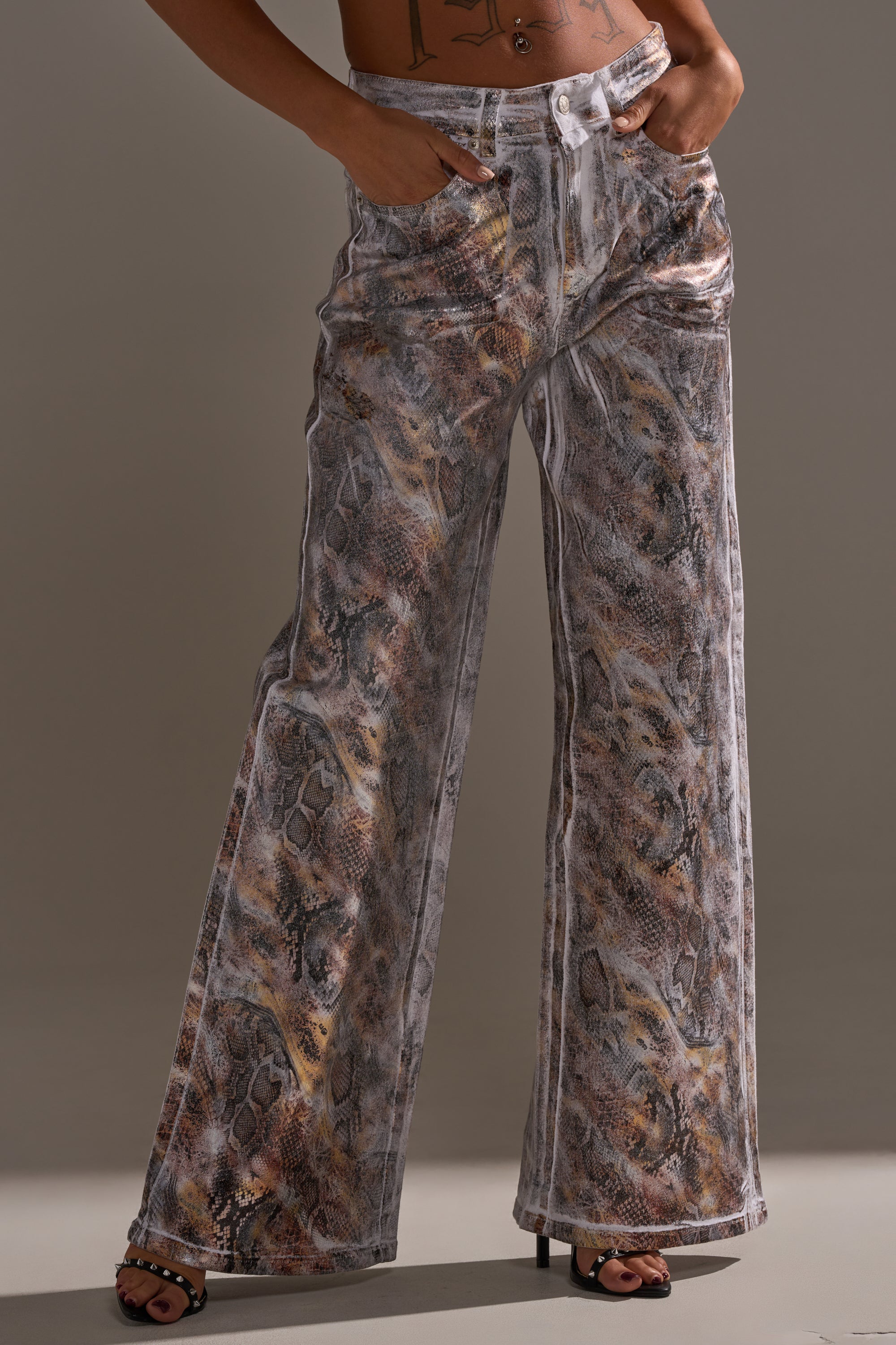A person in the ADDICTED TO YOU METALLIC SNAKE WIDE LEG DENIM—high-rise, wide-leg pants with a gray and brown snake print—stands hands-in-pockets in heeled sandals, tattoo partly visible. Perfect party-ready bottoms.