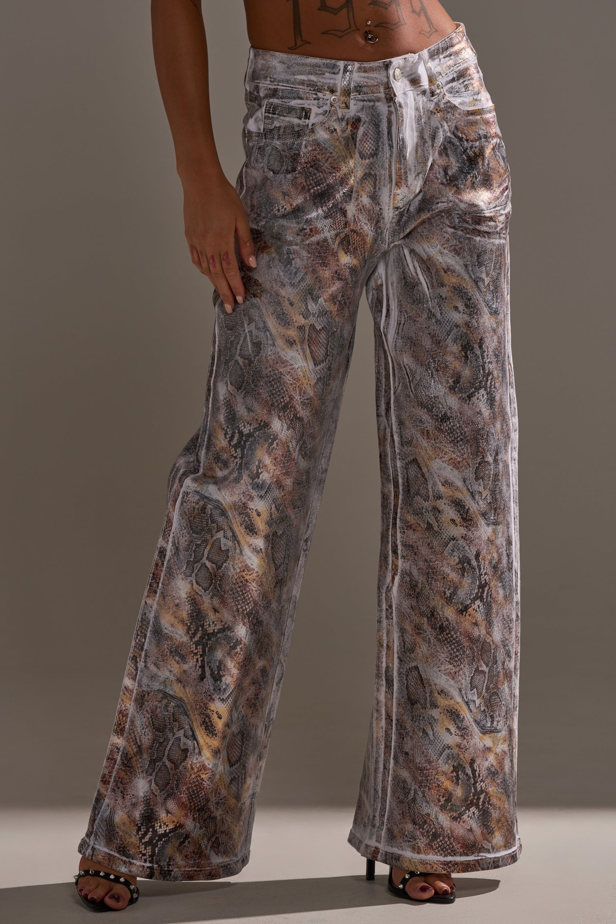 A person models the ADDICTED TO YOU METALLIC SNAKE WIDE LEG DENIM with heeled sandals, striking a party-ready pose against a plain background.