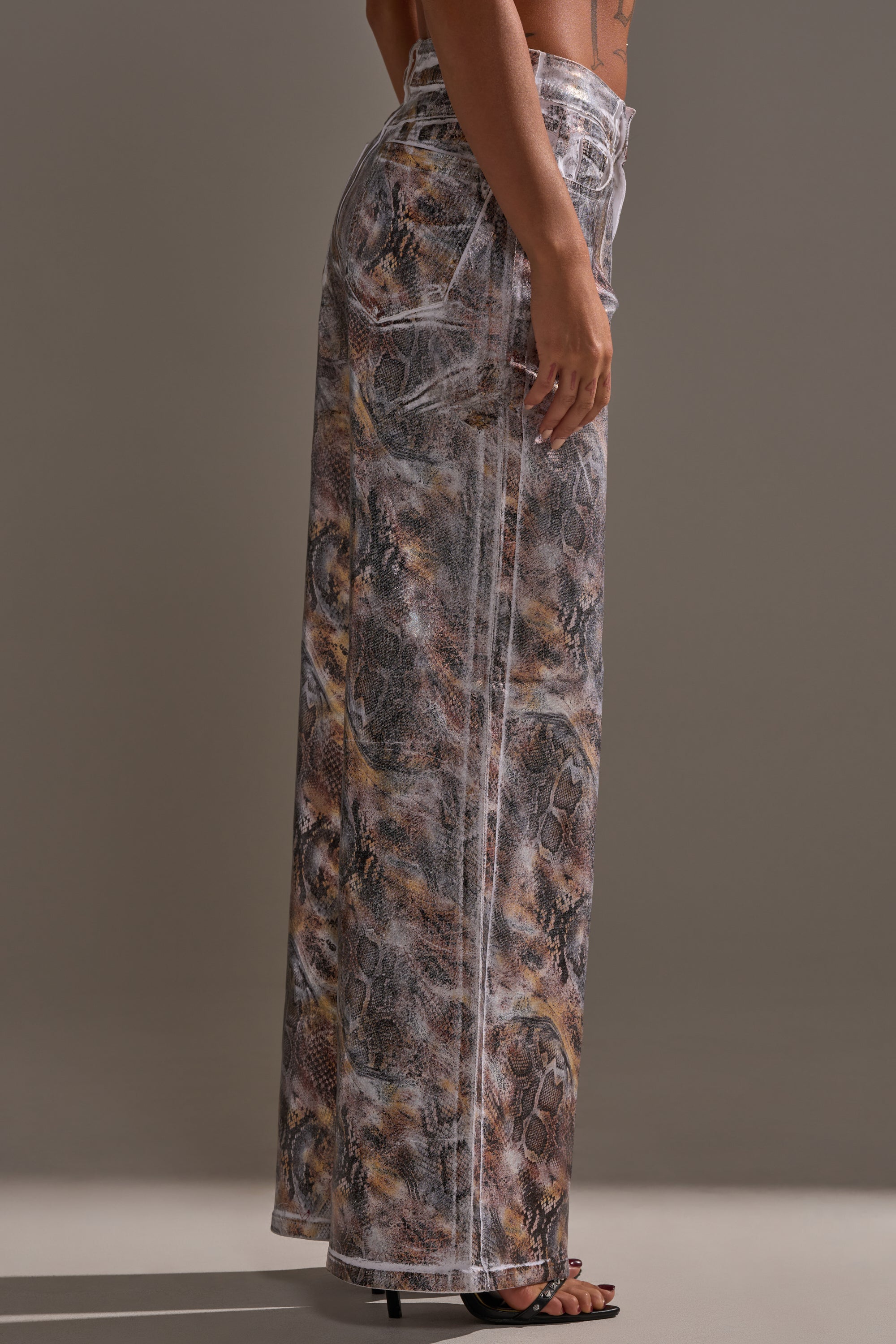A person wears the ADDICTED TO YOU METALLIC SNAKE WIDE LEG DENIM, featuring a high-rise fit and sheer snake print with white seam detailing, shown from the side against a neutral background.