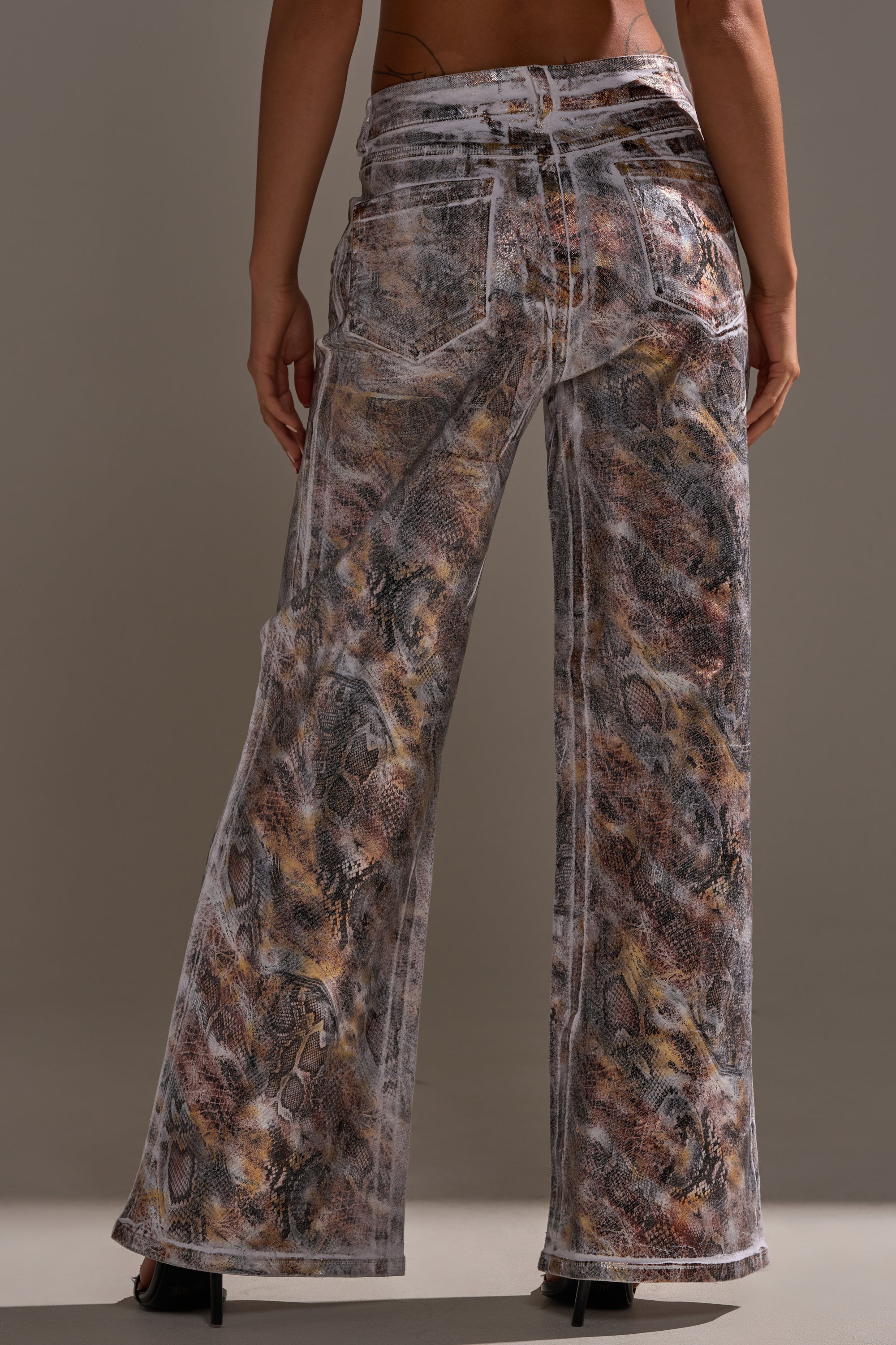 A person stands facing away in ADDICTED TO YOU METALLIC SNAKE WIDE LEG DENIM featuring a high-rise fit, multicolored snakeskin print, white stitching, and black heeled shoes against a plain, neutral background.