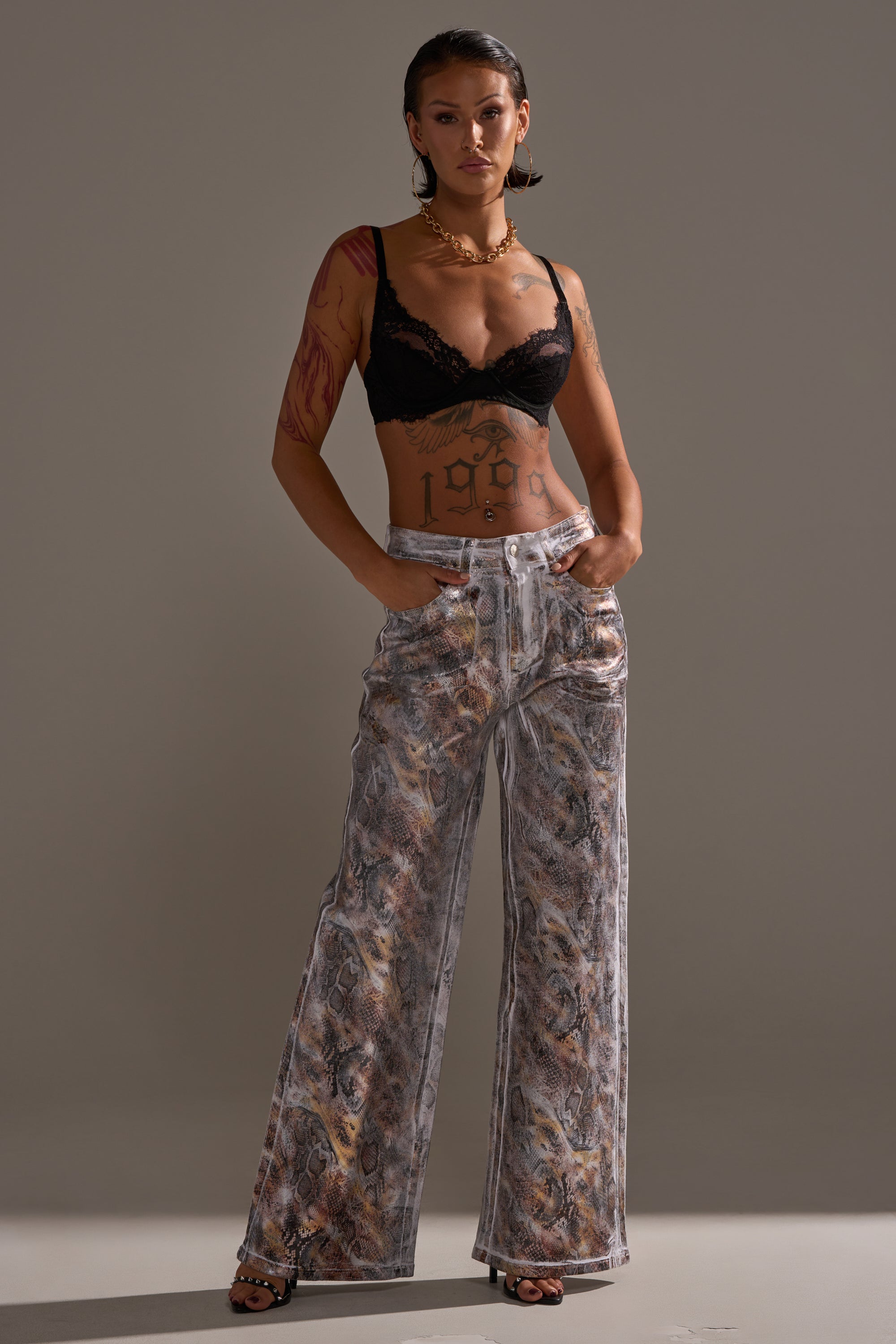 A tattooed woman stands confidently with her hands in her pockets, wearing ADDICTED TO YOU METALLIC SNAKE WIDE LEG DENIM, a black lace bra, and black heels against a plain gray background.