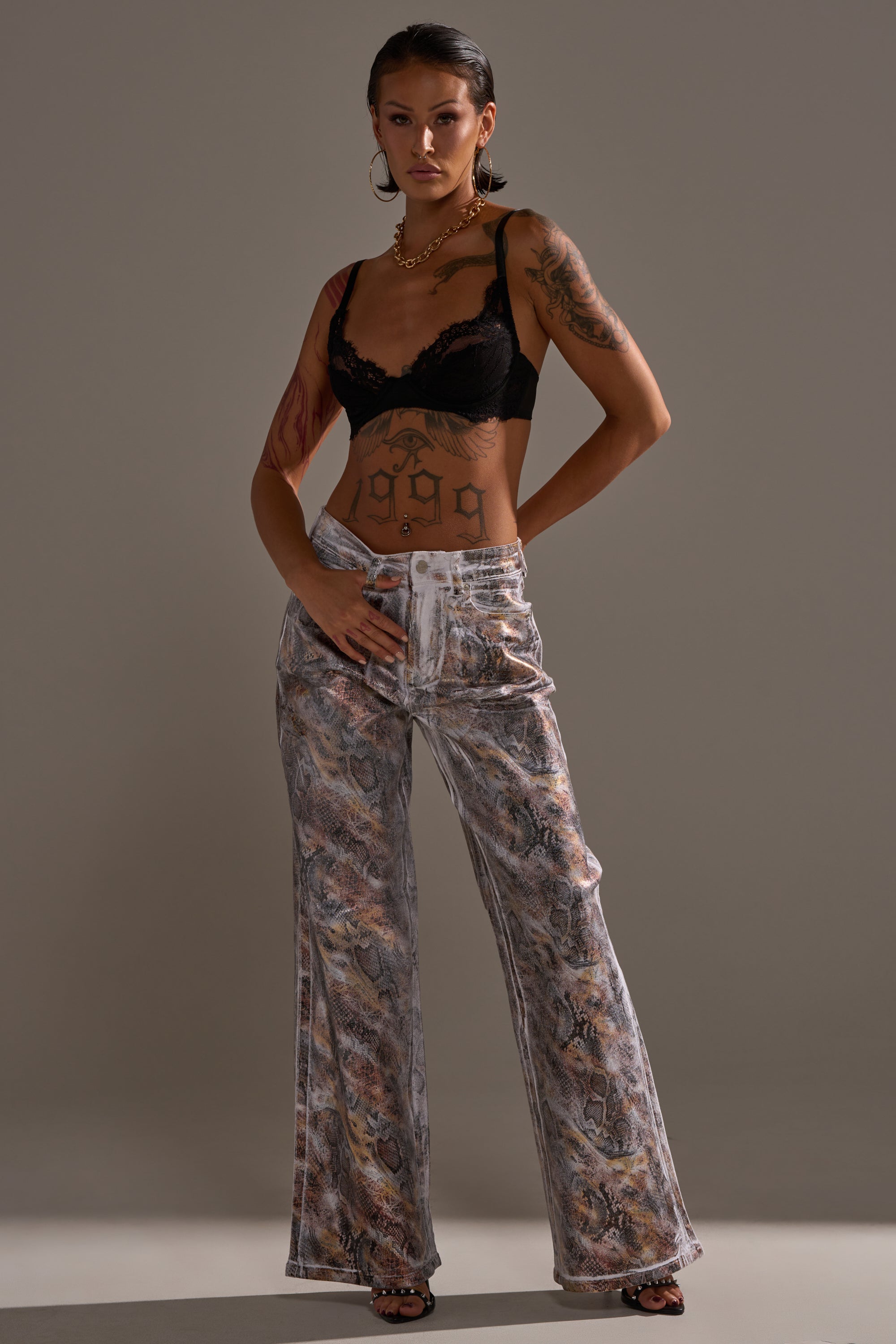 A tattooed woman poses confidently in a black lace bralette and the ADDICTED TO YOU METALLIC SNAKE WIDE LEG DENIM, styled with hoop earrings, a necklace, and open-toed shoes against a plain gray background.