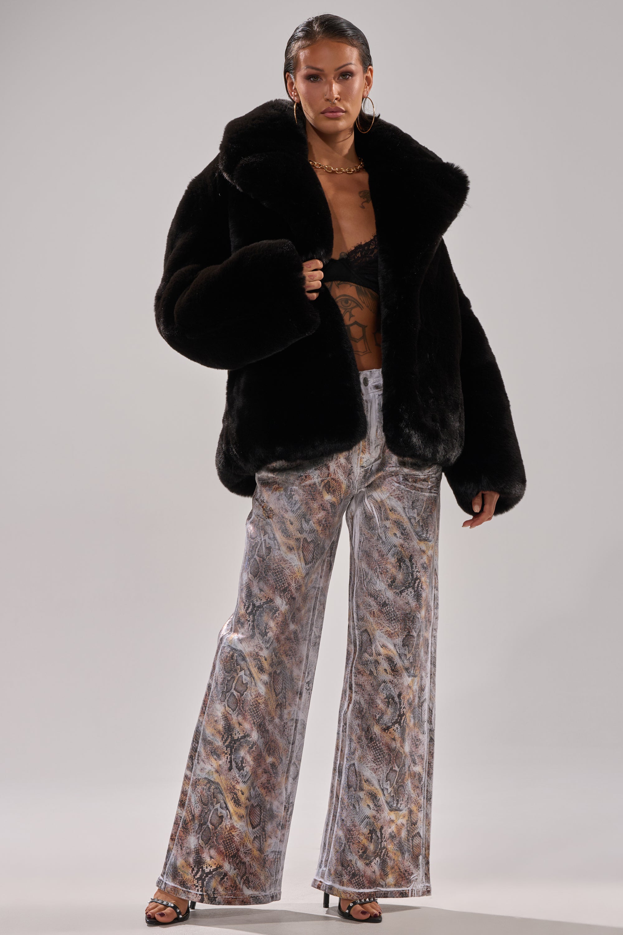 A woman wearing ADDICTED TO YOU METALLIC SNAKE WIDE LEG DENIM makes a bold statement, pairing the standout jeans with a black faux fur coat, black lace bralette, and black open-toe heels against a plain background.