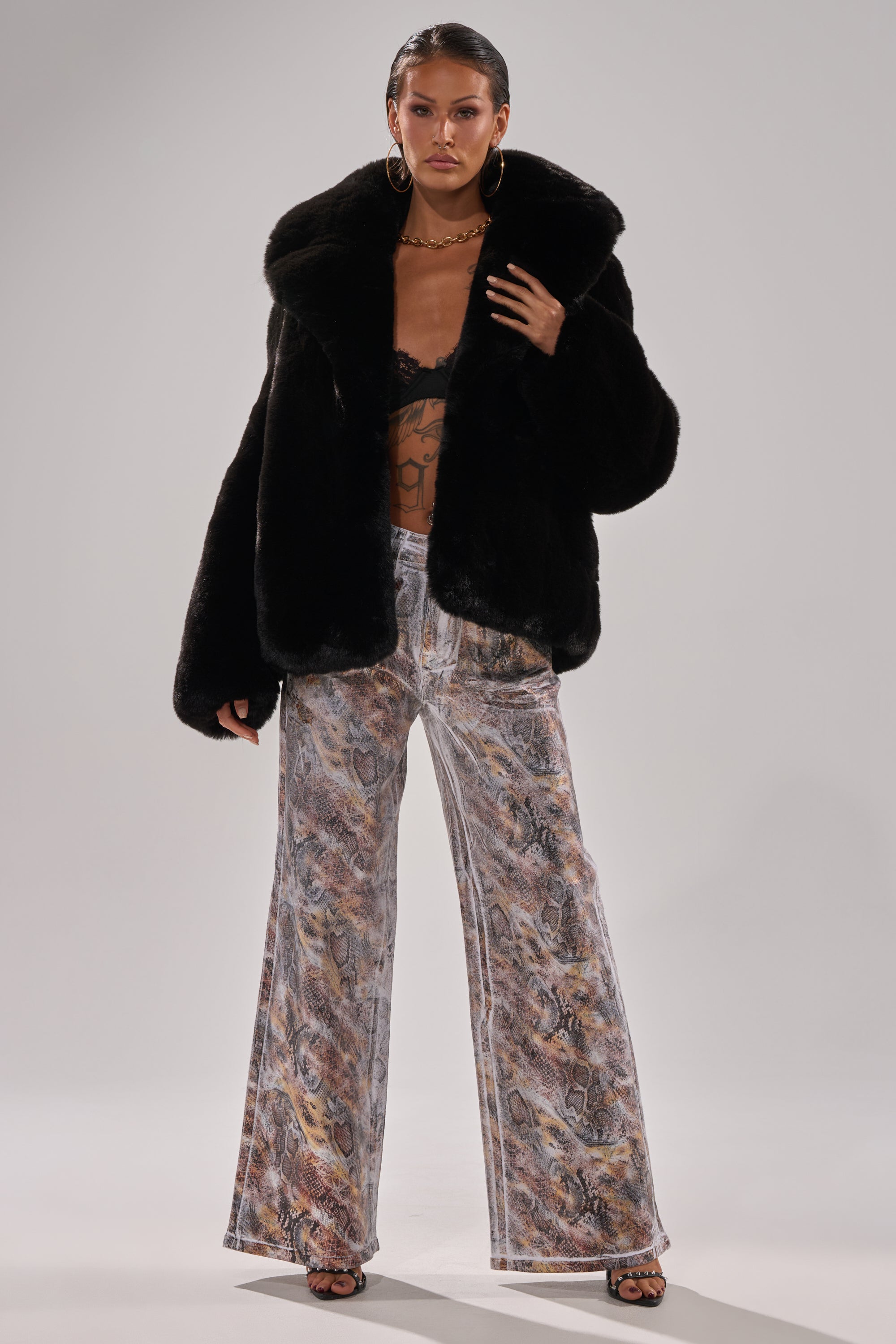 A person poses in black faux fur coat, black lace bralette, ADDICTED TO YOU METALLIC SNAKE WIDE LEG DENIM, large hoop earrings, and open-toe heels against a plain light gray background.