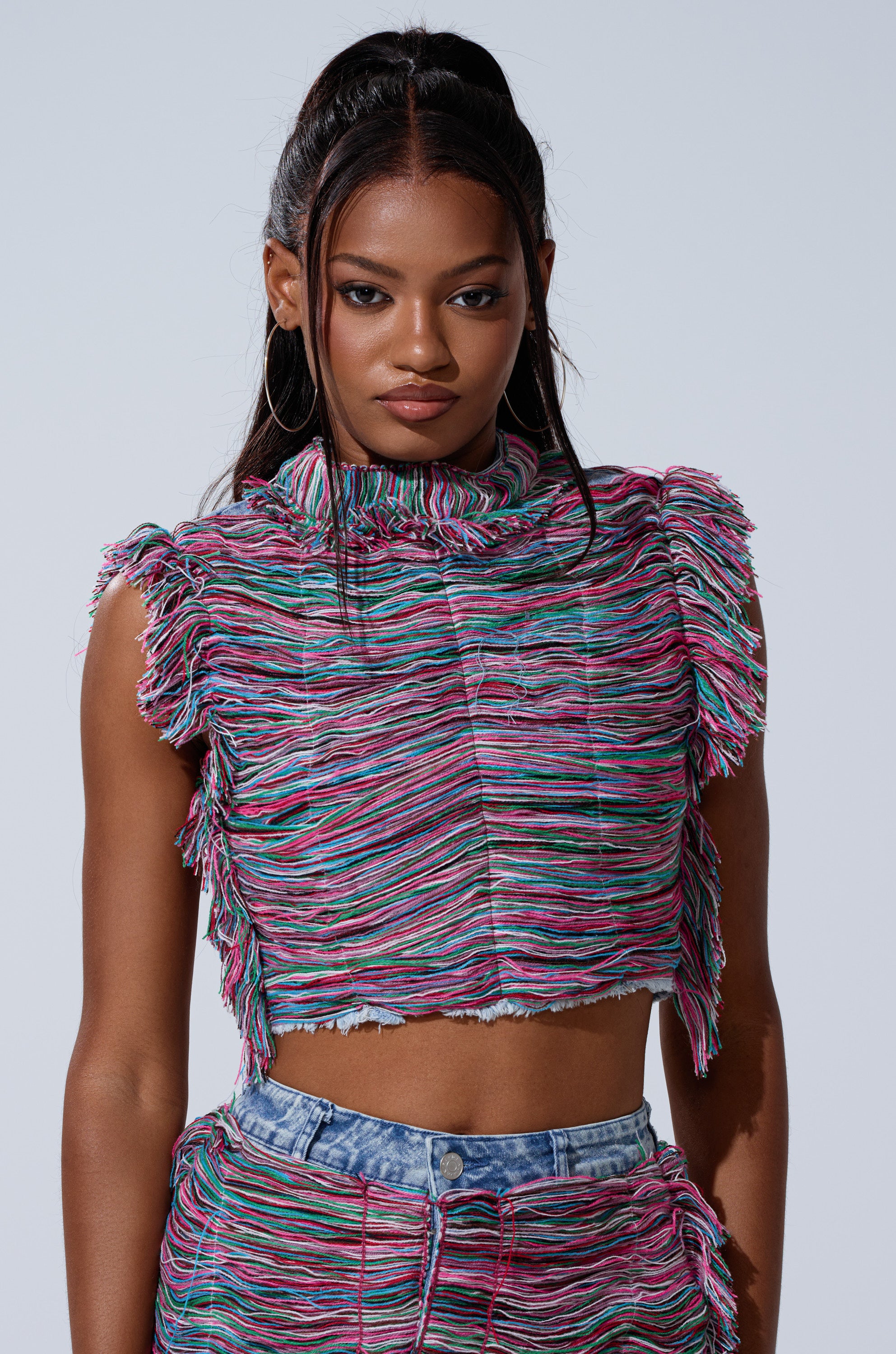 A woman with long dark hair in a high ponytail wears the SHES GONE, DANCE ON GIRL SHREDDED DENIM TOP with high-waisted blue jeans, standing against a plain light background.