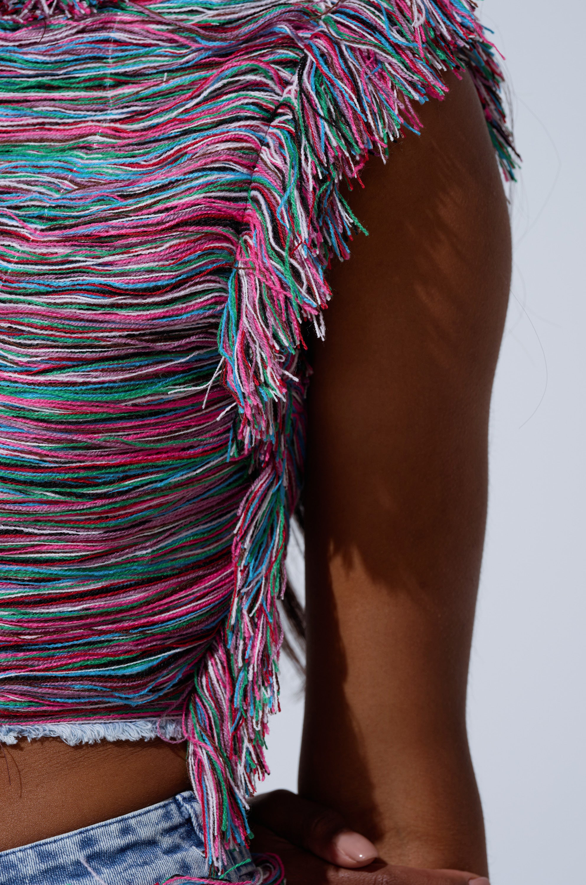 Close-up of a person wearing the SHES GONE, DANCE ON GIRL SHREDDED DENIM TOP, a sleeveless cropped style with pink, blue, and white yarn fringe, paired with blue jeans. Only part of the arm and torso are visible.