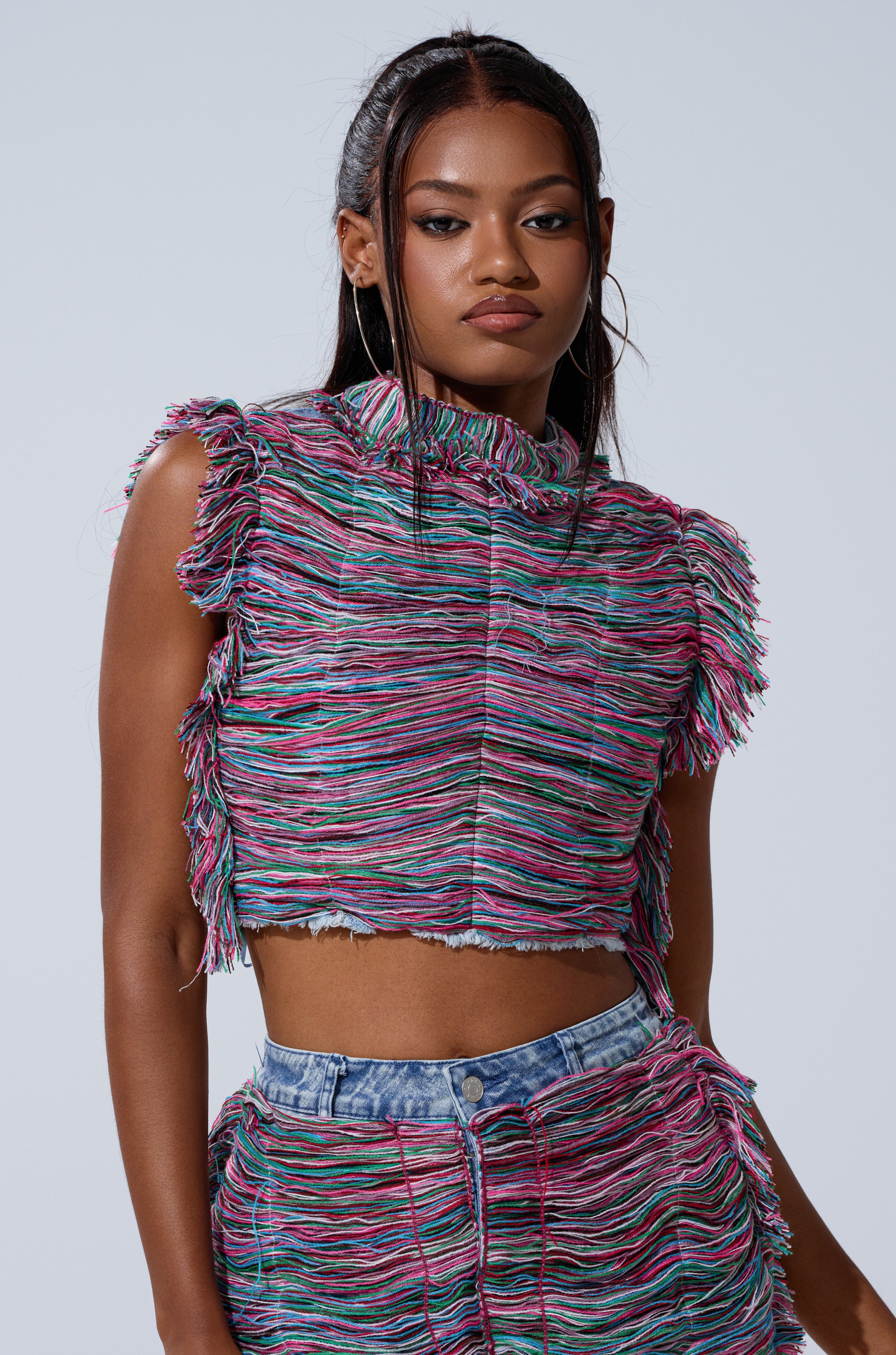 A woman with long dark hair wears the SHES GONE, DANCE ON GIRL SHREDDED DENIM TOP, a colorful textured crop top with multicolored threads and playful yarn fringe. She poses confidently against a plain light background.