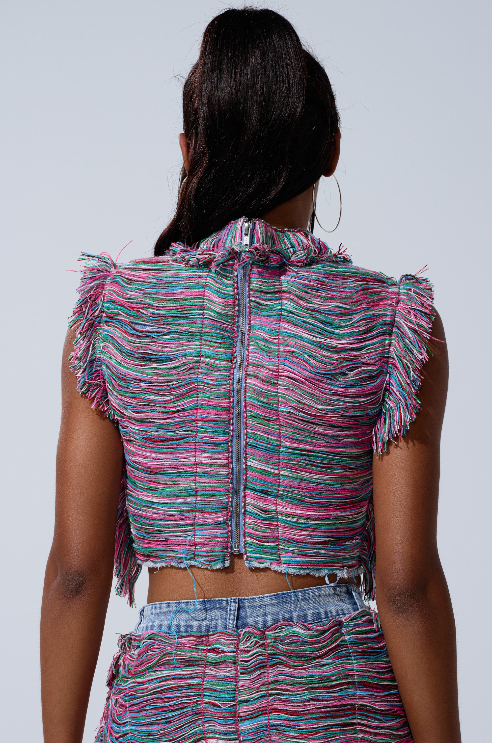 A person with long dark hair in a ponytail is shown from behind against a plain light background, wearing the SHES GONE DANCE ON GIRL SHREDDED DENIM TOP with multicolored yarn fringe and matching bottoms.